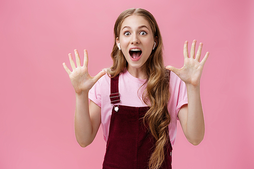 Look no wires. Excited and amazed good-looking charismatic young white woman with wavy natural hair wearing wireless earphones and showing empty raised hands smiling joyful and thrilled over pink wall.