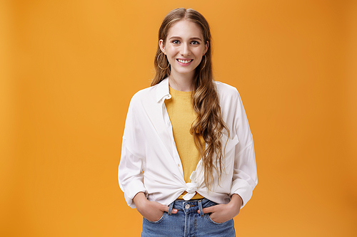 Stylish young female designer wants help friend shopping holding hand in pockets smiling joyfully and self-assured at camera wearing trendy blouse over yellow t-shirt posing over orange wall.