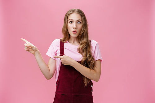 Excited stunned and surprised charming young girl with makeup and curvy hairstyle in trendy corduroy dungarees pointing left folding lips from interest and amazement being curious what happened.