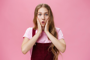 Cute feminine teenage girl in trendy overalls with tattoo on arm folding lips in wow, touching cheeks from amazement and surprise staring at camear shocked and astonished from awesome sales.