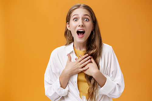 Waist-up shot of excited and thrilled happy expressive european woman in blouse over t-shirt gasping from amazement and surprise holding hands on breast astonished posing over orange background.