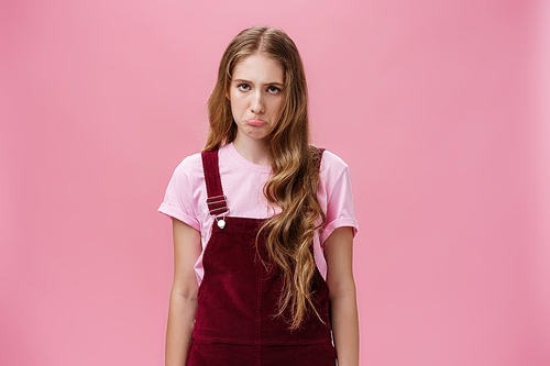 Portrait of silly displeased and uneasy cute young woman in overalls pouting and sulking making gloomy face losing or having bad day standing upset and sad over pink background. Emotions concept