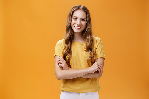 Portrait of kind and friendly charming young female student in casual t-shirt with wavy natural long hair holding hands crossed on chest smiling broadly and carefree at camera over orange background.