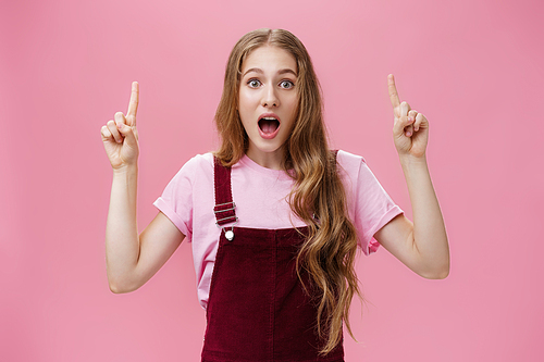 Cute active and charismatic young woman with wavy hair, small scar and tattoo on arm open mouth with amazement and excitement lifting hands to point up at awesome impressive copy space over pink wall. Advertising concept.
