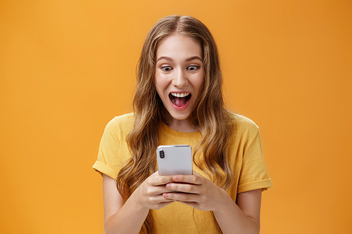 Excited and thrilled charismatic young happy girl in yellow t-shirt smiling with opened from joy mouth holding smartphone staring at cellphone screen astonished carried away with cool phone game.