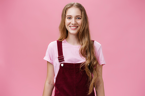 Waist-up shot of kind friendly-looking pleasant young female student with long wavy fair hair in trendy overalls wearing makeup smiling joyfully and gazing at camera with good mood over pink wall.