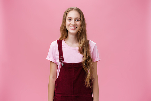 Portrait of charming slim young girl with long fair hair in corduroy overalls smiling joyfully standing upright with positive grin posing against pink background happy and friendly.