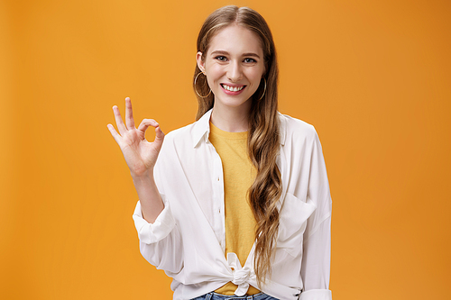 It is okay. Friendly-looking pleasant smiling girl with wavy hair in trendy blouse over t-shirt showing ok or perfect gesture smiling assertive and assured giving positive answer over orange wall.