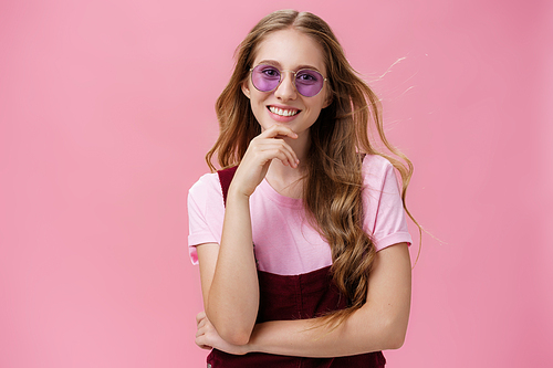 Beauty, style and fashion concept. Portrait of charming confident european female with natural wavy hair in sunglasses and overalls touching chin amused and smiling broadly against pink wall.