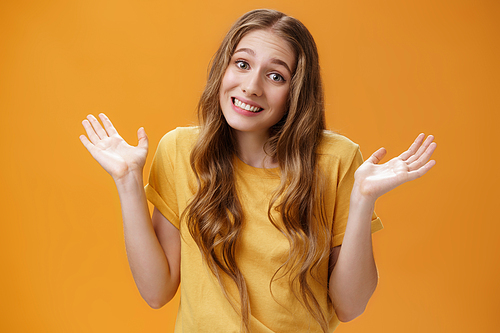 Girl stay out of business shrugging with hands raised and spread aside, silly sorry smile posing over orange background unaware and confused in casual t-shirt having no idea about topic.