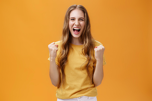 Portrait of happy cheerful lucky young european girl with long natural wavy hair raising clenched fists in cheer and joy yelling yes from triumph feeling glorious moment of win over orange wall. Copy space