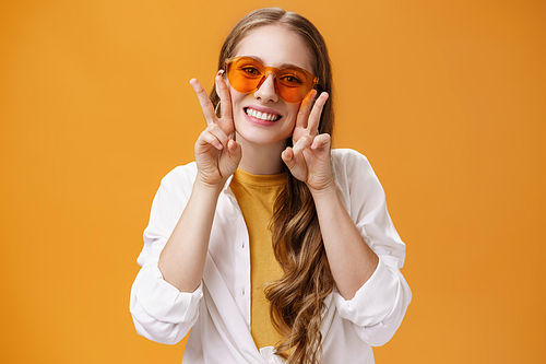 Stylish enthusiastic and charismatic young party girl in trendy sunglasses and white blouse over t-shirt showing peace gestures near face and smiling cute at camera against orange background. Copy space