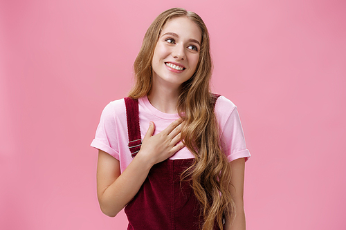 Waist-up shot of delighted charming friendly-looking tender female with cute white smile in corduroy overalls holding palm on breast and gazing right thankful, being grateful for compliments. Body language concept