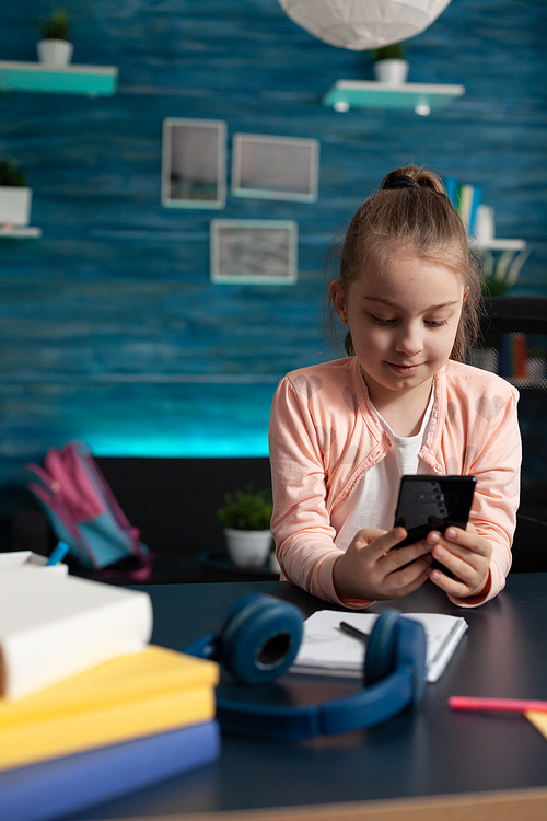 Elementary school pupil holding smartphone at desk for online communication video call conference class. Caucasian little girl using technology for studying lessons and homework