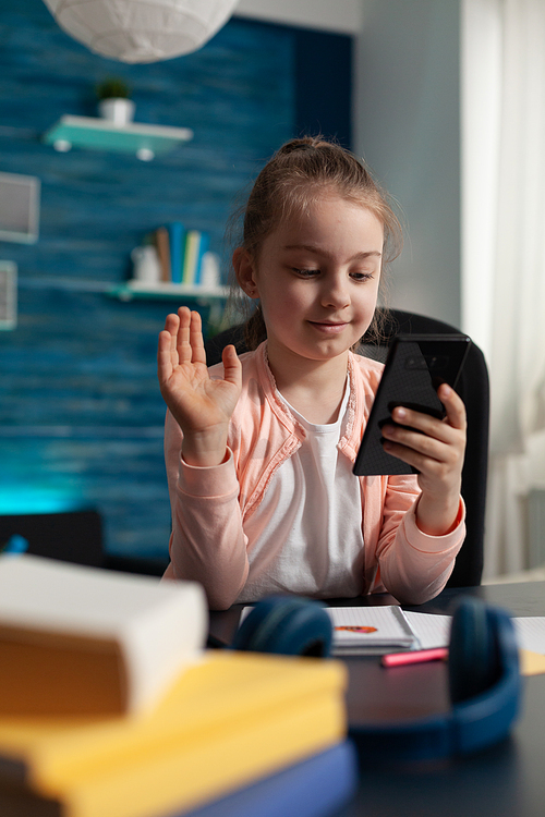 Young student waving at video call camera on smartphone while talking to teacher for online classes and elementary school homework. Small girl sitting at desk indoors with mobile phone