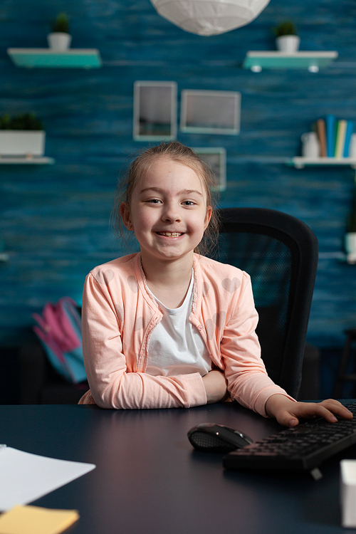 Portrait of little primary school kid sitting at home desk smiling. Caucasian pupil girl waiting for online class lesson on internet to start. Child with notebook on table for homework