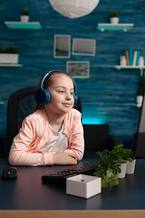 Clever school girl using computer monitor at home while wearing headphone set listening to teacher on online class meeting for education. Small student sitting with technology knowledge