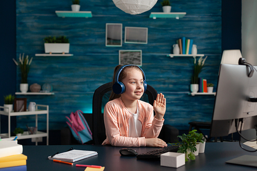 Little student waving at video call conference on computer for online math class using internet technology. Small caucasian girl with headphones and gadget studying for primary education