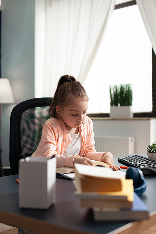 Little schoolgirl taking notes and writing at home desk while attending online class meeting on internet. Elementary school student learning from notebook to pass exam paper for education