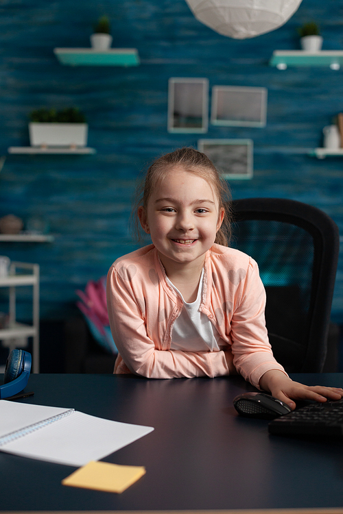 Joyful little girl smiling sitting at desk for online school work on internet classroom lecture. Clever child with educational supplies notebook pen preparing for communication meeting