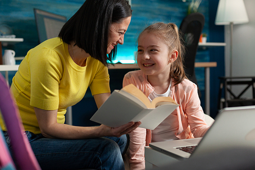Caucasian mother helping girl with homework project at home. Parent assisting child while reading book for education exam and online class lesson lecture with modern laptop at desk