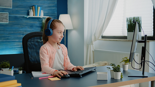 Child waving at video call webcam on computer for school work at desk. Young girl using online conference for class lesson and learning from home. Pupil with headphones on internet