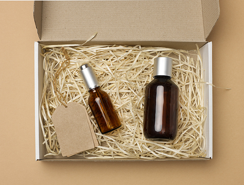 brown glass bottles for cosmetics and paper tags on a rope lie in a cardboard paper box on a beige background, top view