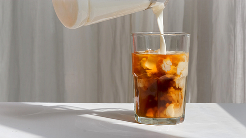 Milk is pouring into a glass with coffee with ice cubes on the background of curtains in the sunlight slow motion