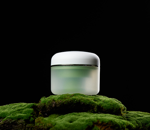 round green plastic jar with white lid for cosmetics stands on green moss, black background. Natural creams and masks. Product branding