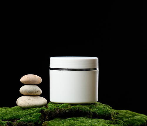 round white plastic jar with lid for cosmetics stands on green moss, black background. Natural creams and masks. Product branding