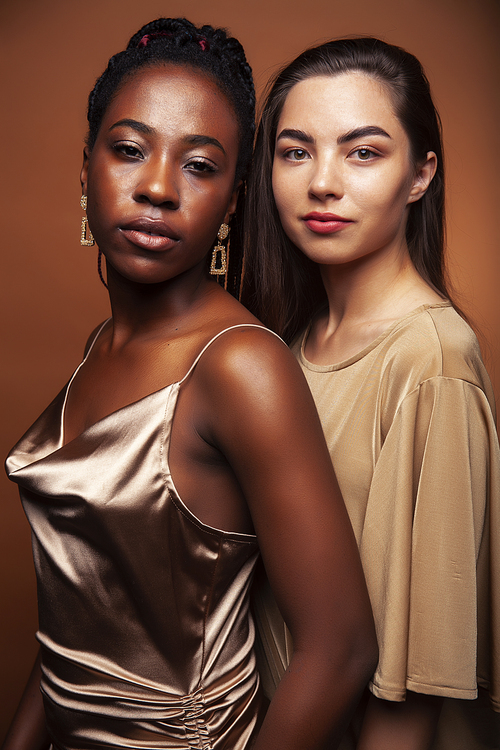 young pretty caucasian, afro woman posing cheerful together on brown background, lifestyle diverse nationality people concept closeup