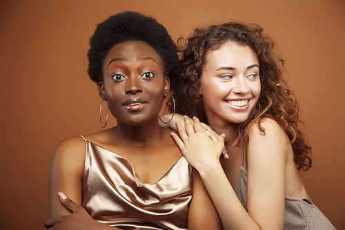 two pretty girls african and caucasian blond posing cheerful together on brown background, ethnicity diverse lifestyle people concept closeup