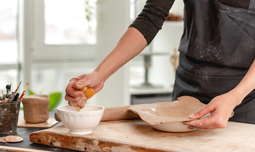 Woman using wet sponge for plate forming from clay at pottery workshop