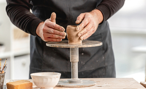 Man using pottery wheel for molding cup from clay at workshop