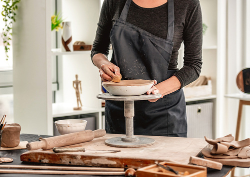 Woman using wet sponge for plate forming from clay on pottery wheel at workshop