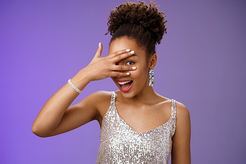 Close-up stylish african american woman afro hairstyle brilliant accessorise glittering silver dress hiding face peeking through fingers amused look surprised anticipating interesting gift.