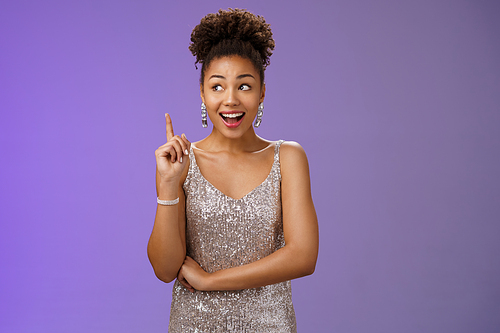 Creative joyful dreamy young african-american girl in silver dress raise index finger eureka gesture look aside inspired have awesome idea sharing suggestion tell plan aloud, standing blue background.