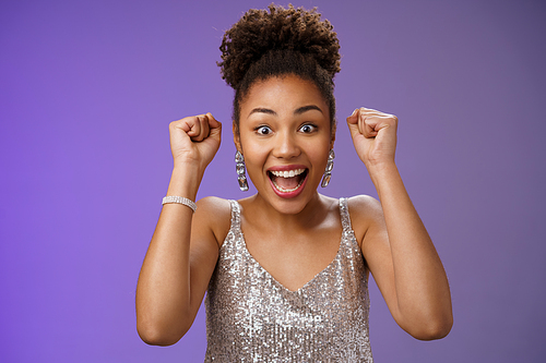 Happy astonished african american woman in silver fashionable dress winning first prize gladly celebrating raising clenched fists dancing triumphing happily having fun, standing blue background.