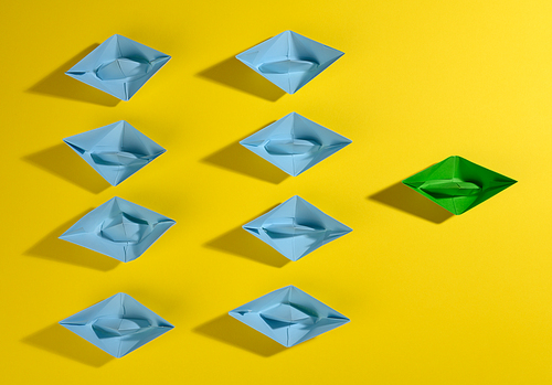a group of blue paper boats and one green led on a yellow background. The concept of a strong leader, manipulation of the masses, a close-knit team and effective management