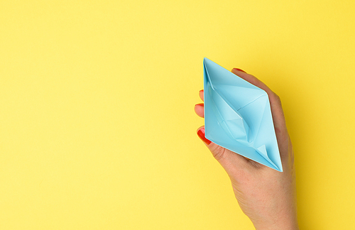 female hand hold a blue paper boat on a yellow background. Mentoring and support concept