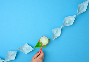 female hand holds a red magnifying glass over a row of paper boats on a blue background. Talent search concept, recruiting, recruitment interviews, top view