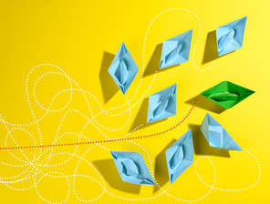 a group of blue paper boats and one green with paths on a yellow background. The concept of a strong leader with extraordinary thinking, quick decision-making. Finding the optimal and simple solution in business