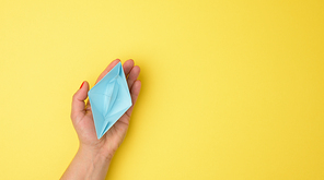 female hand hold a blue paper boat on a yellow background. Mentoring and support concept, top view