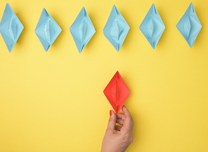 a group of blue paper boats follows a red boat in front of a yellow background. The concept of a strong and charismatic leader in a team, manipulating the masses, top view