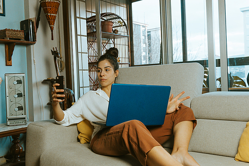Young woman making a call while working on his laptop from home in the couch in the city