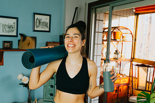 Young woman smiling to camera while holding a bottle of water and a yoga mat at a modern flat in the city during a bright day