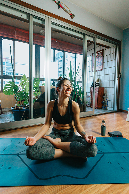 Young woman doing a yoga position over a professional yoga mat while laughing in a modern flat in the city