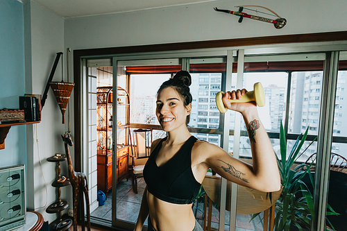 Young woman lifting weights while doing fitness while smiling to camera at a modern and minimalistic flat