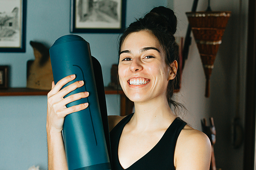 Young woman smiling to camera while holding a yoga mat at a modern and bright flat