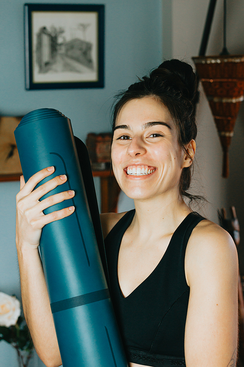 Young woman smiling to camera while holding a yoga mat at a modern and bright flat